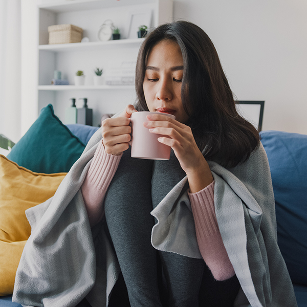 Woman sitting on a couch huddled under a blanket and drinking a hot beverage